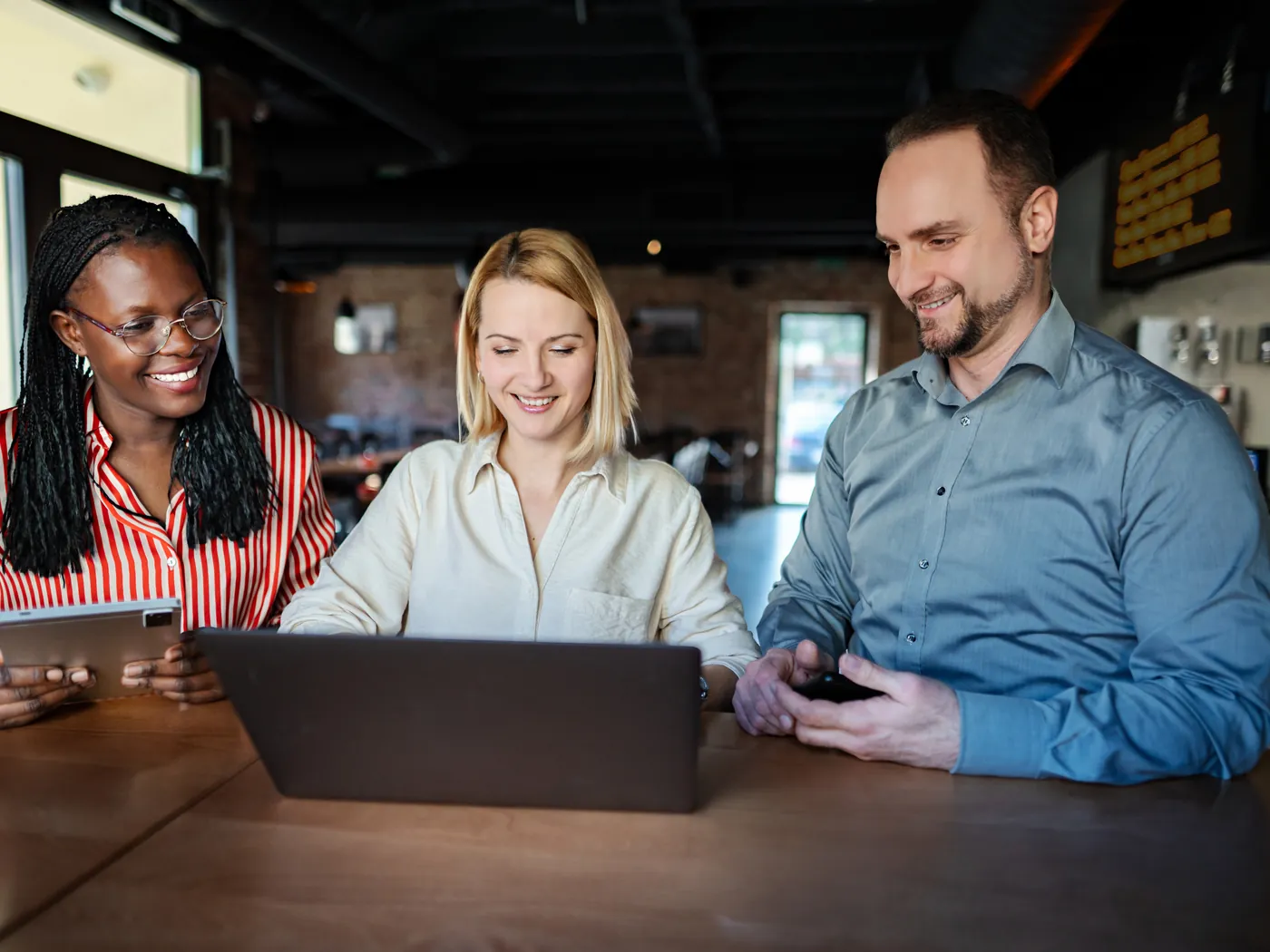 Drie collega's kijken lachend samen naar een laptop aan een houten tafel.