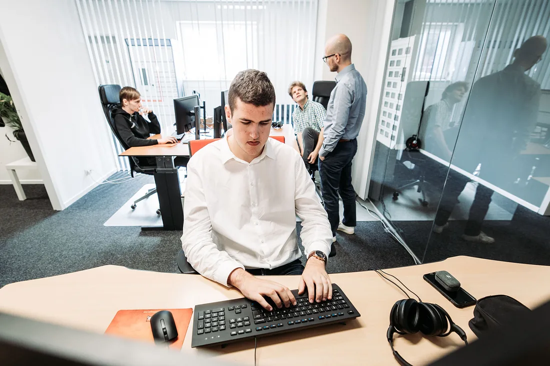 Een engineer van Infodatek Group aan zijn bureau, met collega's in overleg op de achtergrond.