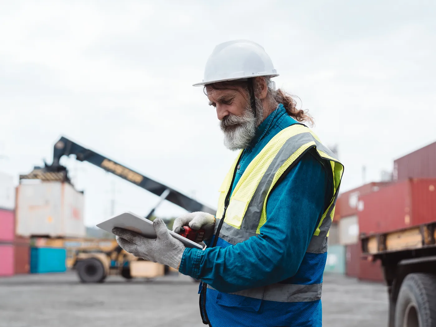 Een medewerker met helm controleert een tablet op een containerterminal, met een reachstacker op de achtergrond.