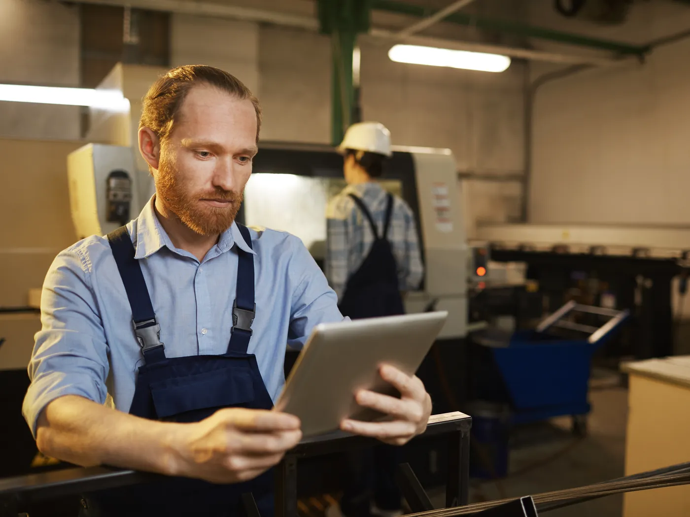 Een monteur in een werkoverall kijkt op een tablet in een productiehal met machines.
