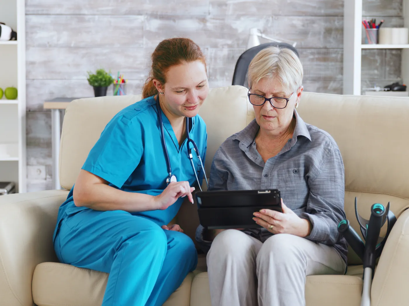 Een zorgmedewerker laat een oudere vrouw op de bank iets zien op een tablet.